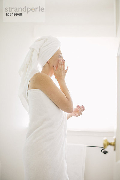 In ein weißes Handtuch gewickelte Frau  die in einem Badezimmer steht und ihr Gesicht eincremt.