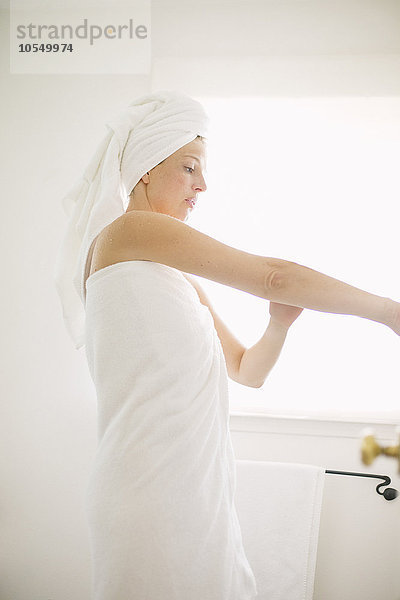 In ein weißes Handtuch gewickelte Frau steht in einem Badezimmer und trägt Lotion auf ihren Arm auf.