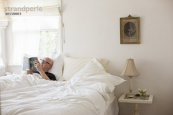 Mann liegt in einem Bett mit weißer Bettwäsche und liest.