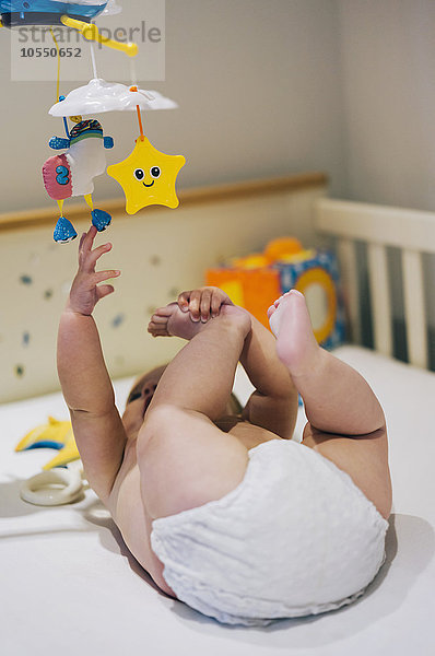 Ein kleines Mädchen in einer Windel  das in einem Kinderbett liegt und bis zu einem bunten Mobile reicht  das über ihm hängt.