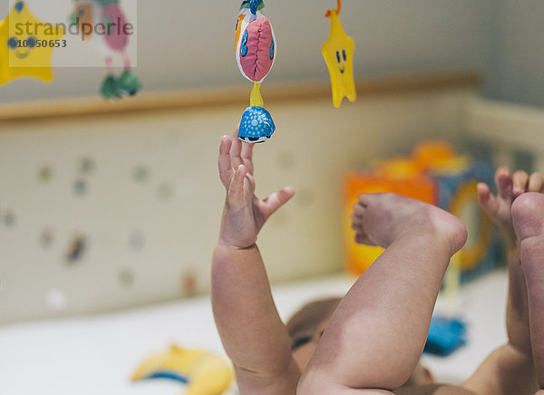 Ein kleines Mädchen in einer Windel  das in einem Kinderbett liegt und bis zu einem bunten Mobile reicht  das über ihm hängt.