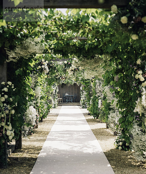 Ein Gartenweg durch einen Bogen führt zu einer Nische mit Tisch und Stühlen  mit Kletterpflanzen und weißen Rosen.