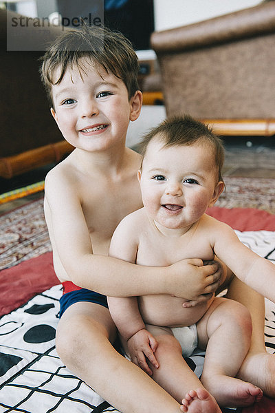 Zwei Geschwister  ein Junge und seine kleine Schwester sitzen lachend auf einem Teppich.