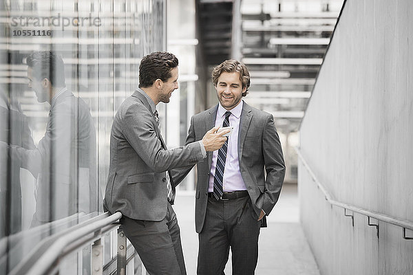 Zwei Männer in Geschäftsanzügen vor einem großen Gebäude  einer mit einem Smartphone in der Hand.