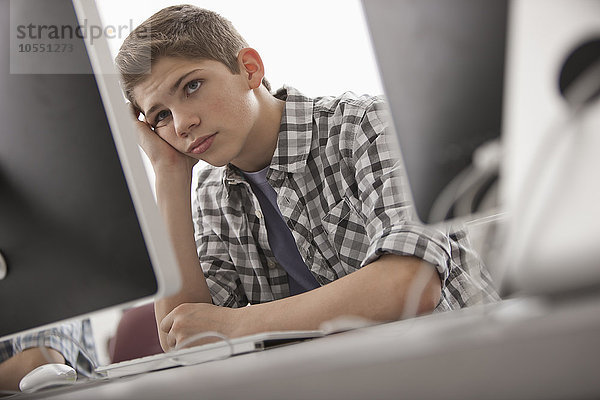 Ein Schulraum  Computerlabor oder Labor mit Reihen von Computermonitoren und Sitzgelegenheiten. Ein Junge  der mit der Hand am Kinn sitzt und die Nase voll hat.