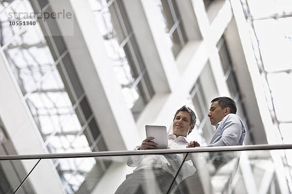 Zwei Männer  Geschäftskollegen  stehen an einem Geländer in einem Atrium oder Innenhof und schauen auf ein digitales Tablet.