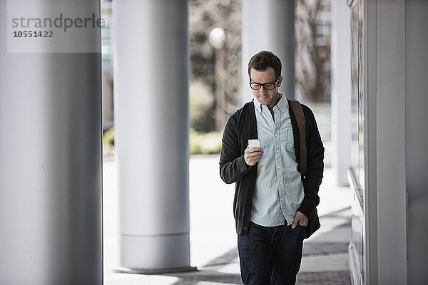 Ein Mann in legerer Kleidung steht vor einem Gebäude und überprüft sein Smartphone.