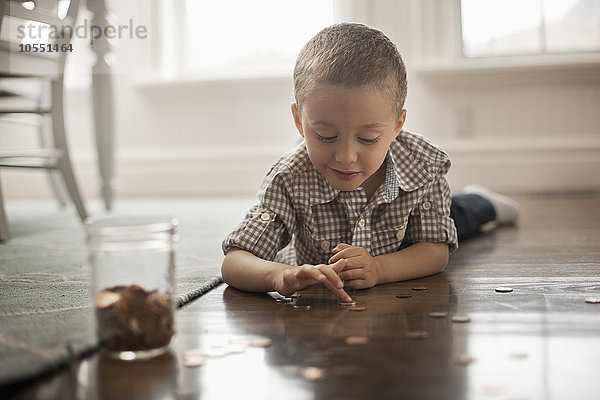 Ein Kind  das auf dem Bauch auf dem Boden liegt  mit Münzen spielt und sie in ein Glasgefäß legt.