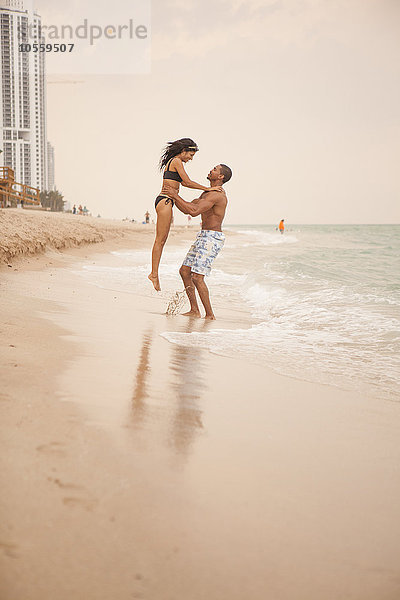 Mann hebt Freundin am Strand in die Wellen des Ozeans
