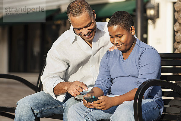 Afroamerikanischer Vater und Sohn benutzen ein Handy auf einer Bank