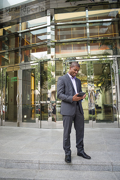 Schwarzer Geschäftsmann benutzt digitales Tablet vor einem Bürogebäude