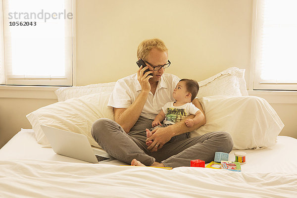 Vater hält seinen Sohn und spricht auf dem Bett mit dem Handy