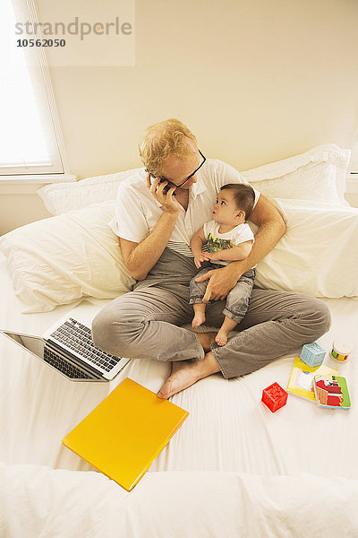 Vater hält seinen Sohn und spricht mit dem Handy auf dem Bett