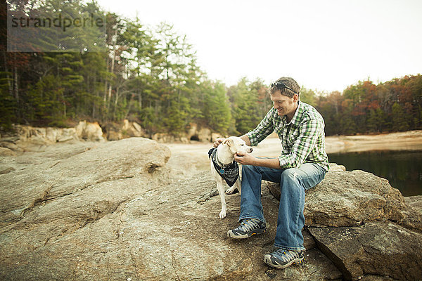 Mann streichelt Hund auf Felsen am See