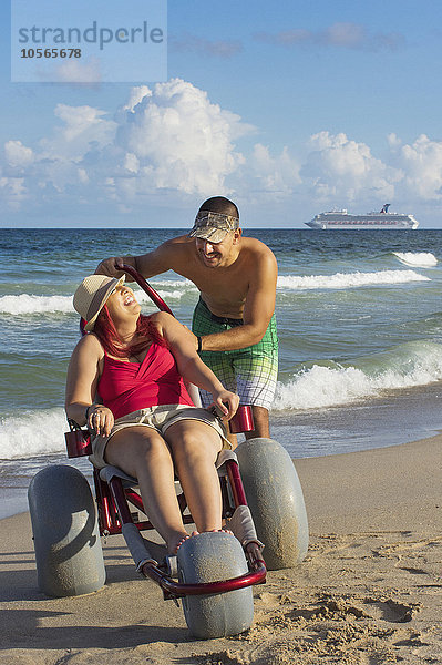 Mann schiebt querschnittsgelähmte Freundin im Rollstuhl über den Strand