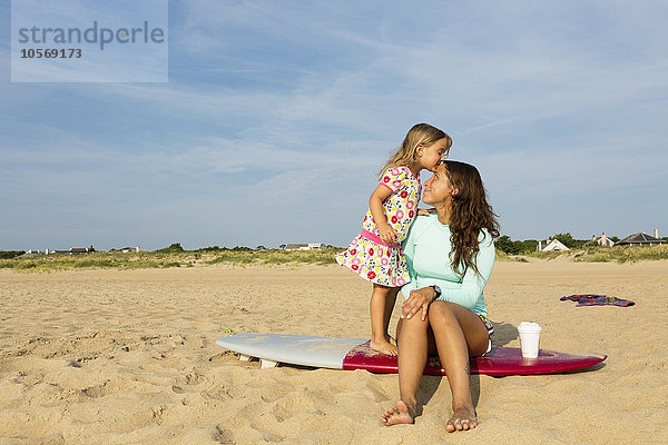 Mädchen küsst Stirn der Mutter auf Surfbrett am Strand