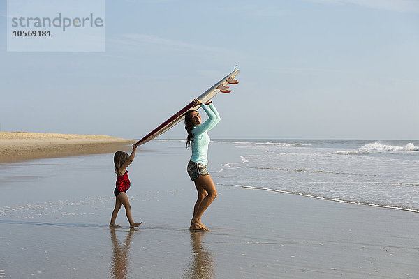 Mutter und Tochter tragen Surfbrett am Strand