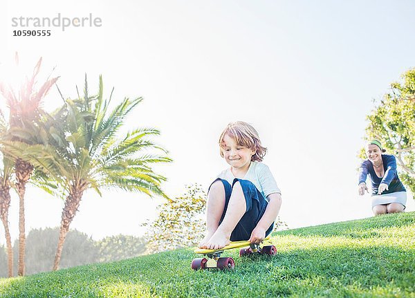 Kleiner Junge sitzt auf dem Skateboard und rollt den Berg hinunter.