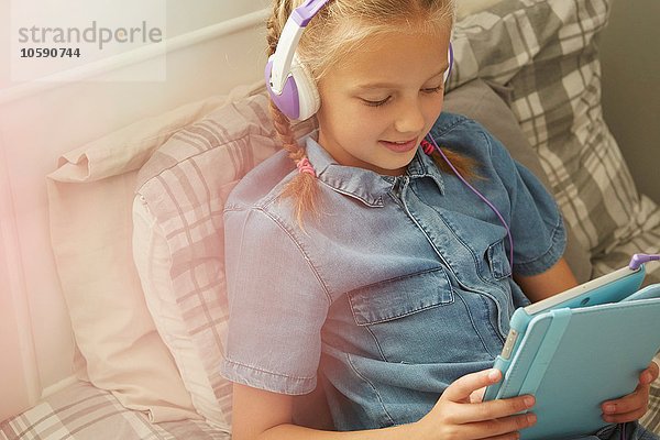 Ausschnitt eines Mädchens  das mit Kopfhörern auf dem Bett sitzt und lächelnd auf ein digitales Tablett blickt.