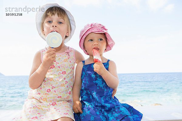 Weibliches Kleinkind und Schwester tragen Sonnenhüte und essen Eis am Strand