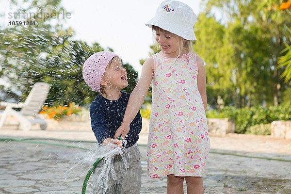 Mädchen und Kleinkindschwester spielen mit Wasserschlauch im Garten