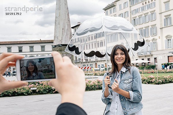 Junge Frau mit Regenschirm  die für einen Freund posiert und mit dem Smartphone fotografiert  Piazza Santa Maria Novella  Florenz  Toskana  Italien