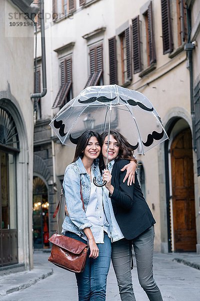 Lesbisches Paar  das zusammen in der Straße steht und den Regenschirm hält und lächelnd in die Kamera schaut  Florenz  Toskana  Italien