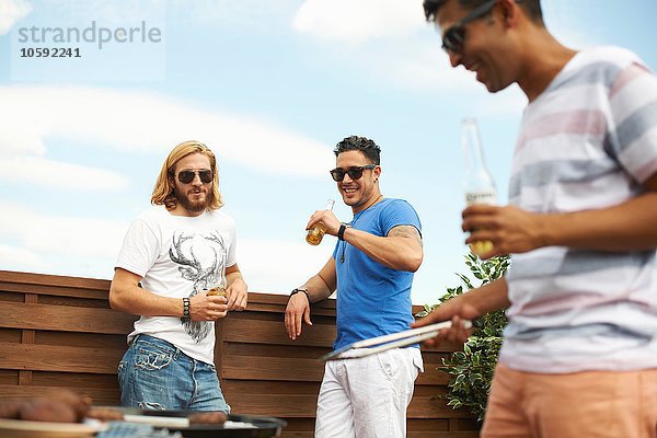 Drei männliche Freunde beim Biertrinken und Grillen auf dem Dach des Hauses