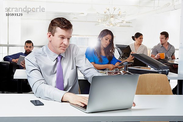 Mittlerer erwachsener Geschäftsmann am Schreibtisch sitzend  mit Laptop  im Hintergrund arbeitende Kollegen
