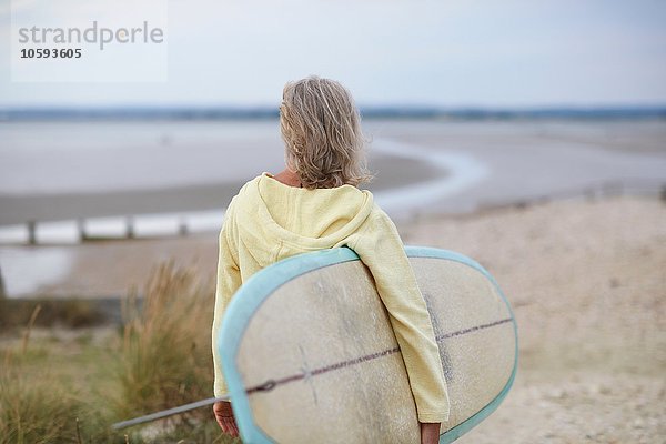 Seniorin auf dem Weg zum Strand  mit Surfbrett  Rückansicht