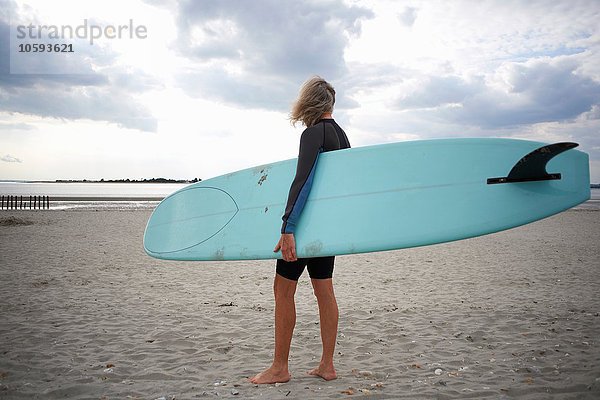 Seniorin steht am Strand  hält Surfbrett  Rückansicht