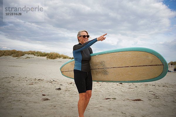 Seniorin steht am Strand  hält Surfbrett  zeigt auf das Meer