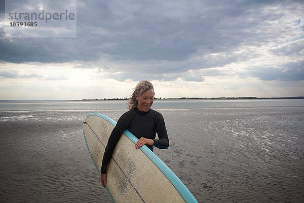 Seniorenfrau  die vom Meer aus geht und Surfbrett trägt