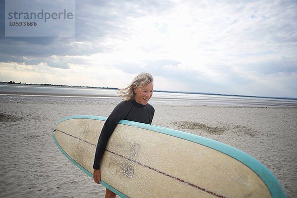 Ältere Frau  die vom Meer geht und Surboard trägt.