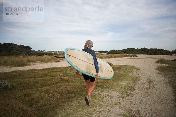 Seniorin  die am Strand spazieren geht  Surfbrett trägt  Rückansicht