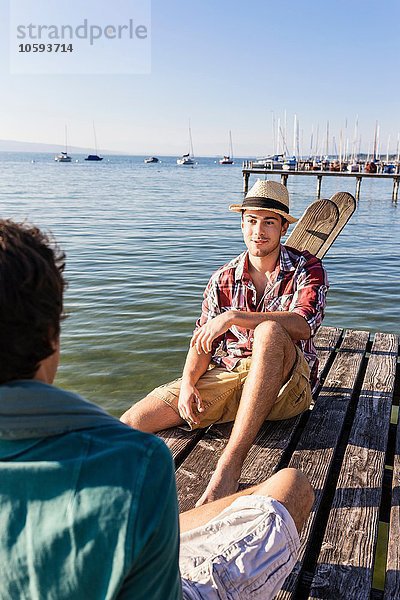 Freunde sitzen von Angesicht zu Angesicht auf einem Holzsteg am See  Schondorf  Ammersee  Bayern  Deutschland