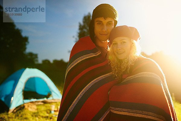 Porträt eines jungen  romantischen Campingpaares  das bei Sonnenuntergang in eine Decke gehüllt ist.