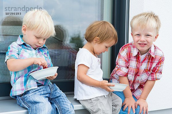 Weibliches Kleinkind und zwei junge Brüder auf der Terrasse  die Schalen mit Himbeeren essen.