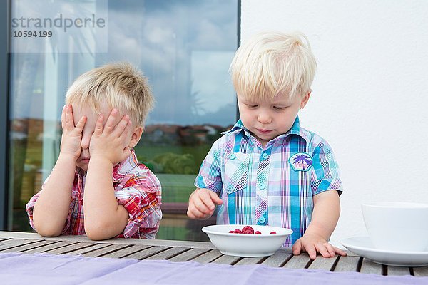 Junge mit deckenden Augen  während der kleine Bruder eine Schale Himbeeren isst.