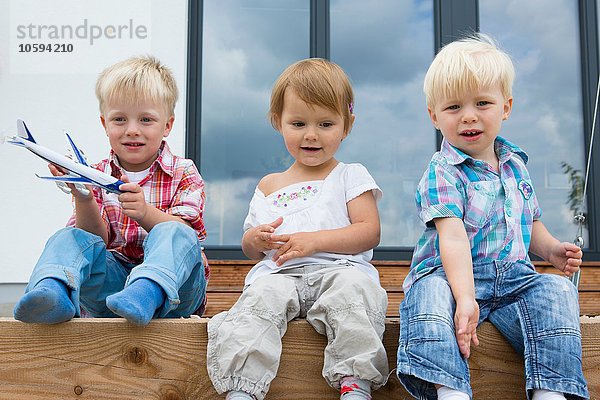 Junge mit Spielzeugflugzeug und zwei Kleinkindern auf der Terrasse sitzend