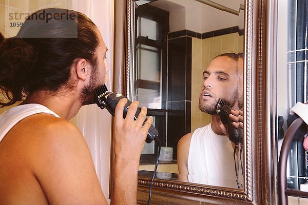 Junger Mann schaut in den Spiegel und rasiert sich mit einem elektrischen Rasierapparat.