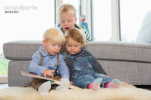 Junge und zwei Kleinkinder lesen Kinderbuch auf dem Wohnzimmerboden