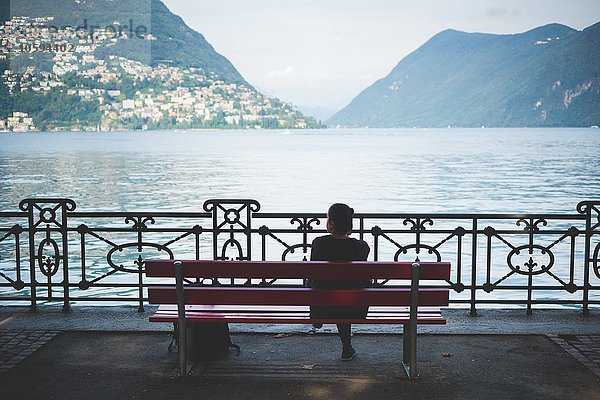 Rückansicht der Silhouette einer Frau auf einer Parkbank mit Blick auf den Luganer See.