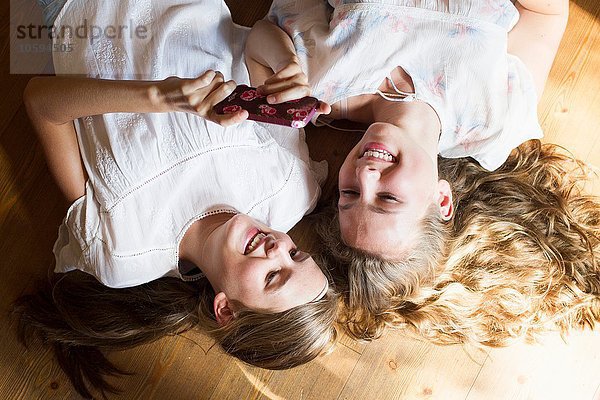 Zwei Teenager-Mädchen liegen auf dem Holzboden und lesen Smartphone-Texte.