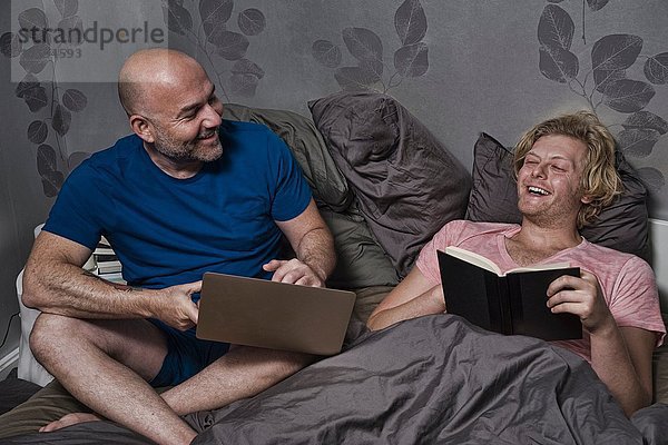 Männliches Paar sitzt im Bett und lacht beim Lesen und Benutzen des Laptops.