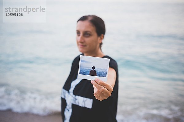 Frau hält ein Foto von sich selbst am Strand hoch.
