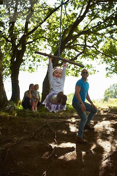 Junges Mädchen schwingt auf Baumschaukel  während ihre Familie zuschaut.