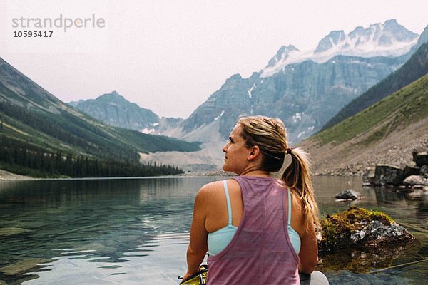 Rückansicht der mittleren erwachsenen Frau  die am Wasser sitzt und wegschaut  Moraine Lake  Banff National Park  Alberta Canada