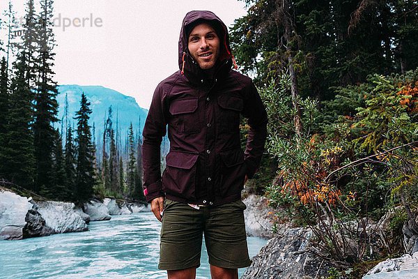 Mittlerer Erwachsener Mann am Fluss mit wasserdichtem Kapuzenmantel  der lächelnd auf die Kamera schaut  Moraine Lake  Banff National Park  Alberta Canada