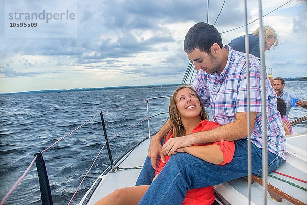 Junge Frau sitzt zwischen den Beinen junger Männer auf einem Segelboot und lächelt von Angesicht zu Angesicht.
